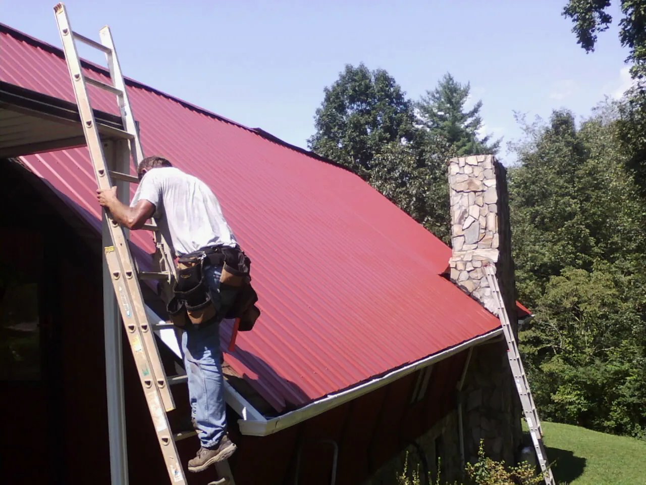 Roofer on a ladder beside a red metal roof and stone chimney in Fayetteville
